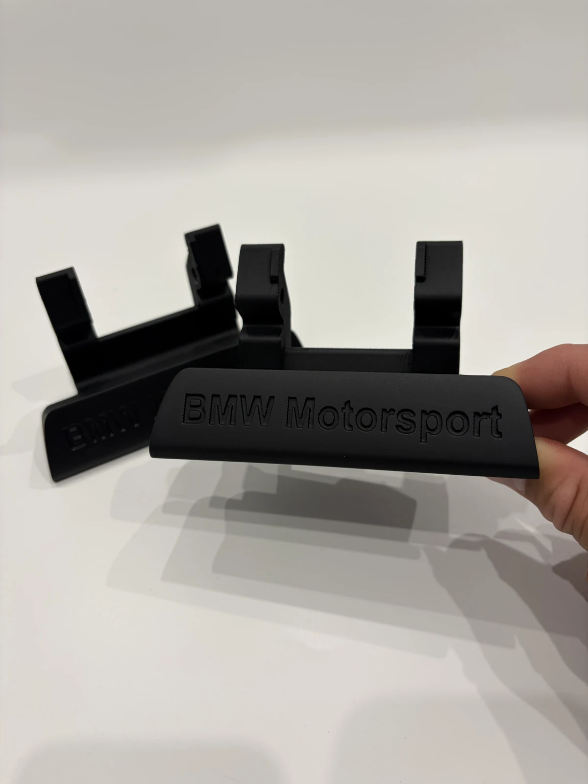 3D spausdinti juodi laikikliai su užrašu BMW Motorsport, laikomi rankoje