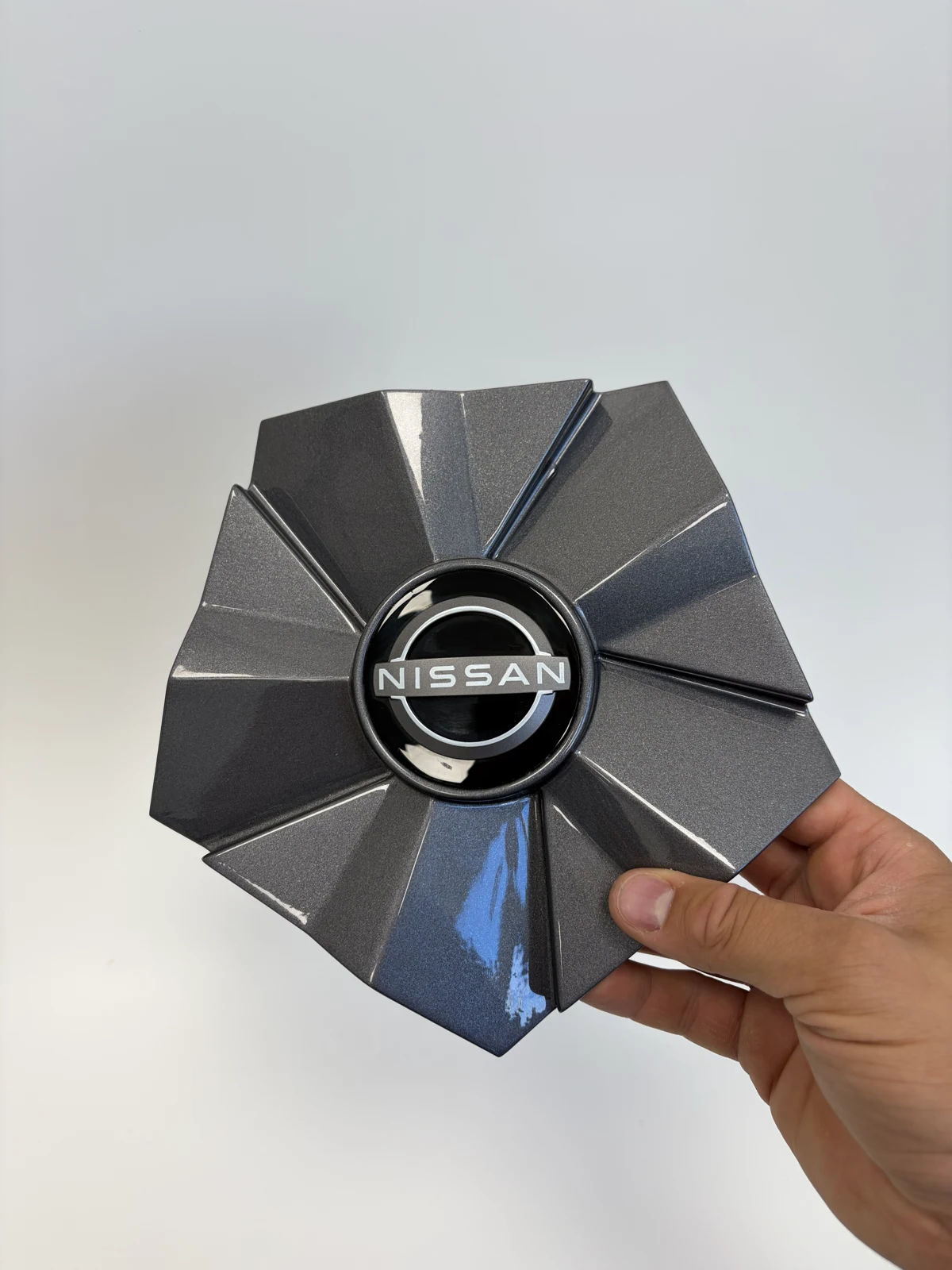 3D spausdintas pilkas blizgus ratlankio dangtelis su Nissan logotipu, laikomas rankoje